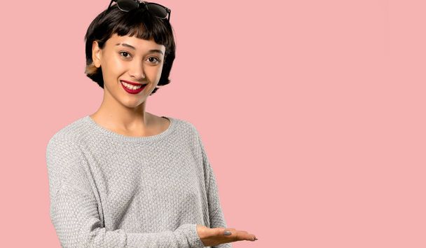 Νεαρή γυναίκα με κοντά μαλλιά παρουσιάζει μια ιδέα ενώ κοιτάζουν χαμογελώντας απέναντι στην απομονωμένη σε ροζ φόντο - Φωτογραφία, εικόνα