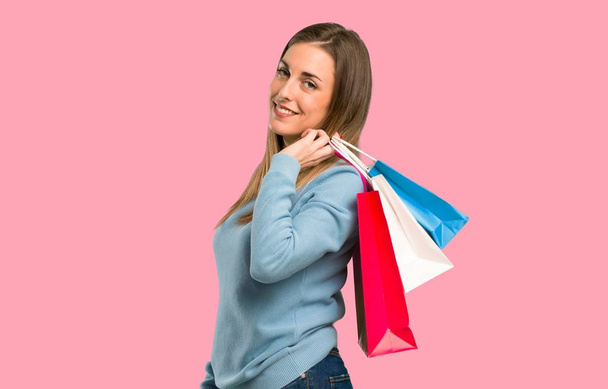 Blonde woman with blue shirt holding a lot of shopping bags on isolated pink background - 写真・画像