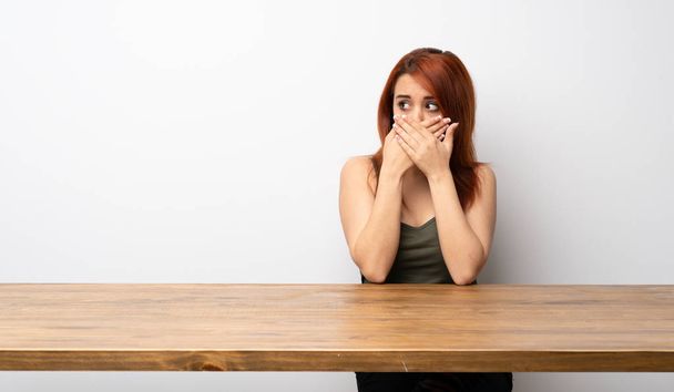 Молодая рыжая женщина за столом закрывает рот и смотрит в сторону
 - Фото, изображение