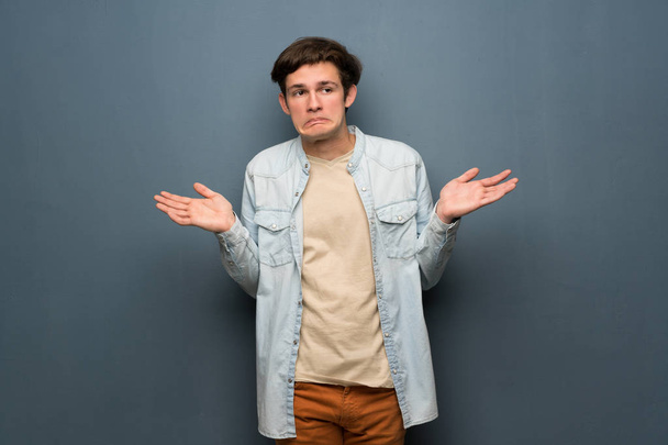 Teenager man with jean jacket over grey wall having doubts while raising hands - Foto, imagen