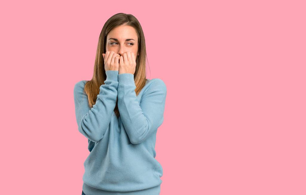 Blonde woman with blue shirt is a little bit nervous and scared putting hands to mouth on isolated pink background - 写真・画像