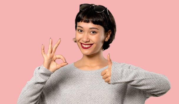 Jonge vrouw met kort haar weergegeven: ok bord met en het geven van een duim omhoog gebaar op geïsoleerde roze achtergrond - Foto, afbeelding