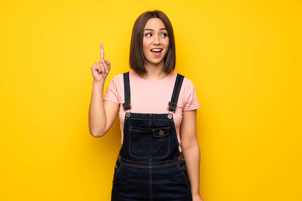 Молода жінка над жовтою стіною має намір зрозуміти рішення, піднімаючи палець вгору
 - Фото, зображення