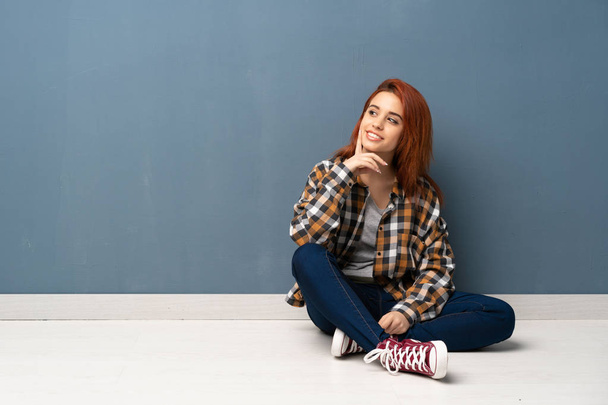 Νέοι κοκκινομάλλα γυναίκα που κάθεται στο πάτωμα σκέψης μια ιδέα ενώ ψάχνει - Φωτογραφία, εικόνα