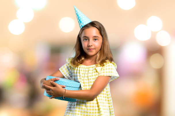 Niña en una fiesta de cumpleaños sosteniendo un regalo manteniendo los brazos cruzados sobre un fondo desenfocado
 - Foto, Imagen