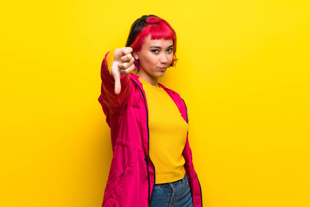 Giovane donna con i capelli rosa sopra parete gialla mostrando pollice verso il basso con espressione negativa
 - Foto, immagini