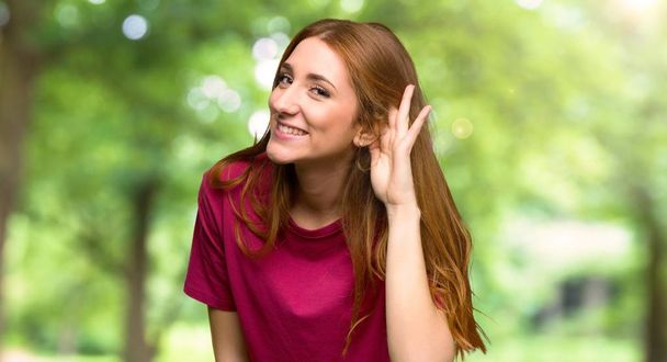 Young redhead girl listening to something by putting hand on the ear in the park - Фото, изображение