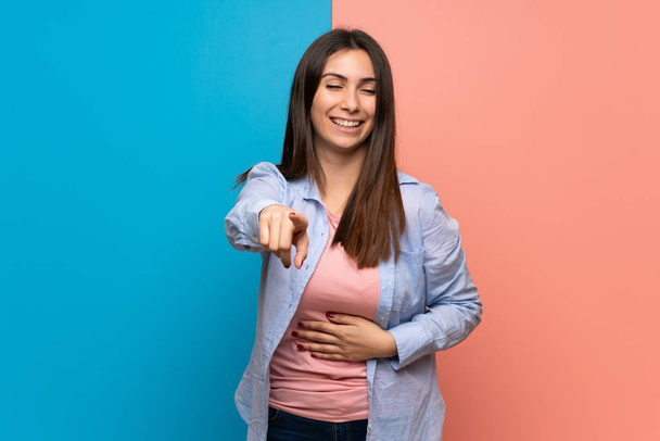 Mujer joven sobre la pared rosa y azul señalando con el dedo a alguien y riendo mucho
 - Foto, Imagen