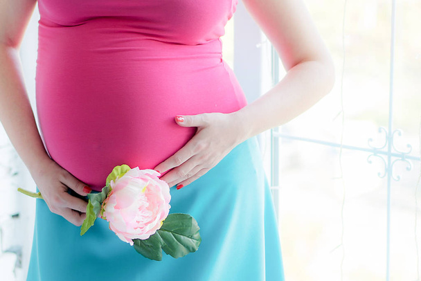 Close-up Image of pregnant woman in nice white dress touching her belly with hands and holding a bouquet of peonies. Beautiful image of a pregnant girl with flowers. Expecting a child. Motherhood. - Photo, image