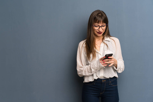 Nainen, jolla on lasit sinisellä seinällä, lähettää viestin matkapuhelimella
 - Valokuva, kuva
