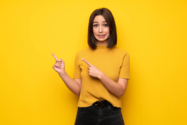 Young woman over yellow wall frightened and pointing to the side - Photo, Image