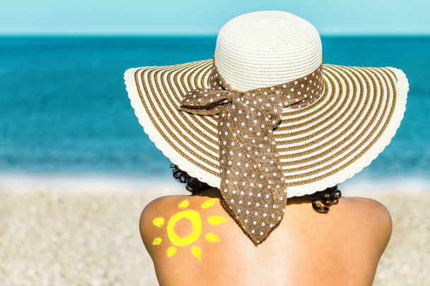 junge Frau mit Hut sitzt am Strand und hat eine Sonne aus Sonnencreme auf der Schulter. - Foto, Bild