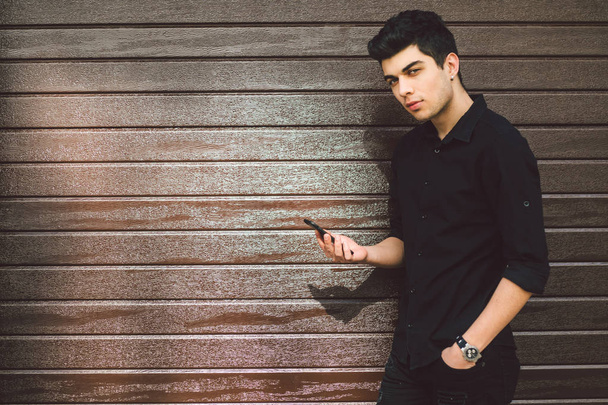 Portrait of young handsome Turkish male model. Mediterranean race brunette in black shirt is using a hand phone of a technology businessman on the street in sunny weather on a wooden wall background - Photo, Image