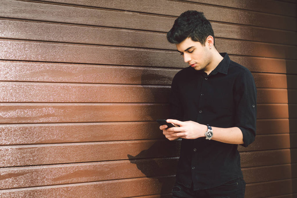Portrait of young handsome Turkish male model. Mediterranean race brunette in black shirt is using a hand phone of a technology businessman on the street in sunny weather on a wooden wall background - Фото, зображення