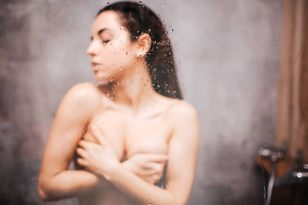 Young attractive sexy woman in shower. Blurred picture. Drk hailred model cover her breast with hands and looking to side. Eyes closed. Posing. - Fotografie, Obrázek