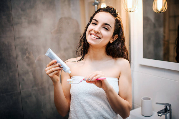Young sporty dark-haired beautiful woman doing morning evening routine at mirror. She look on camera and smile. Putting tooth paste onto tooth brush. - Zdjęcie, obraz