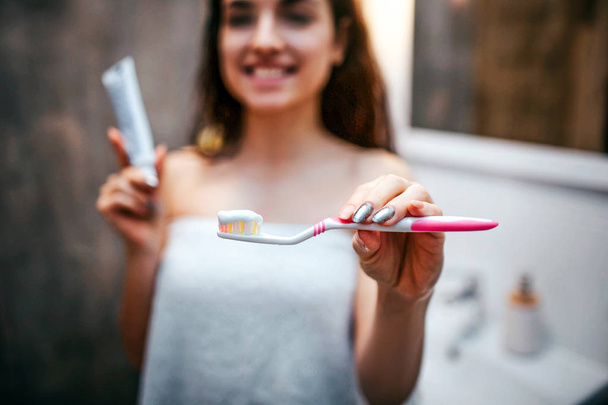 Young sporty dark-haired beautiful woman doing morning evening routine at mirror. Positive nice model show tooth paste with tooth brush closer. She smile. Holding tube of tooth paste. - Φωτογραφία, εικόνα