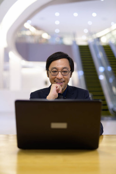 Onnellinen aasialainen liikemies ajattelee käyttäessään kannettavaa toimistorakennuksen sisällä
 - Valokuva, kuva