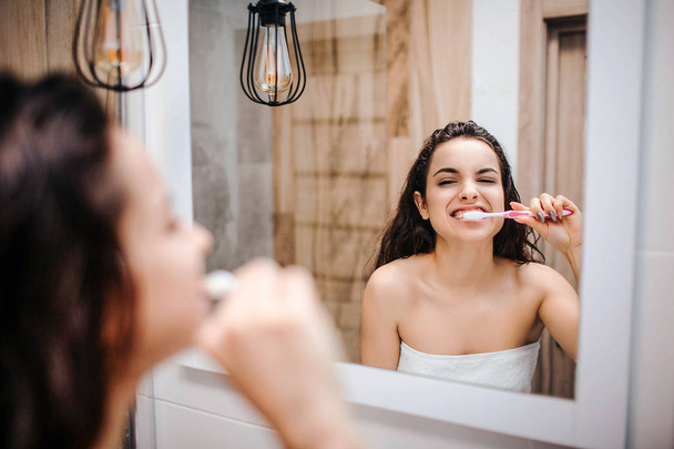 Young sporty dark-haired beautiful woman doing morning evening routine at mirror. She clean her teeth very intense. Model look in the mirror. Body wrapped with white towel. - Photo, Image