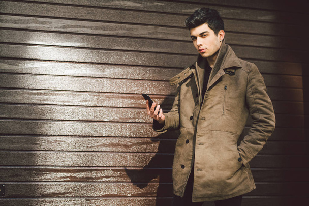 Portrait of young handsome Turkish male model of Mediterranean race brunet in leather coat using hand phone of technology businessman on street in sunny weather against background of wooden wall - Photo, Image