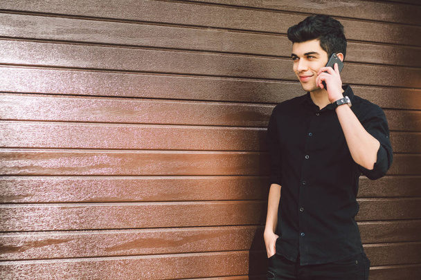 Modelo retrato joven guapo macho sospechoso turco medio oriente morena en camisa negra utiliza teléfono de mano para llamar a la tecnología en la calle en tiempo soleado contra el fondo de una pared de madera
 - Foto, imagen