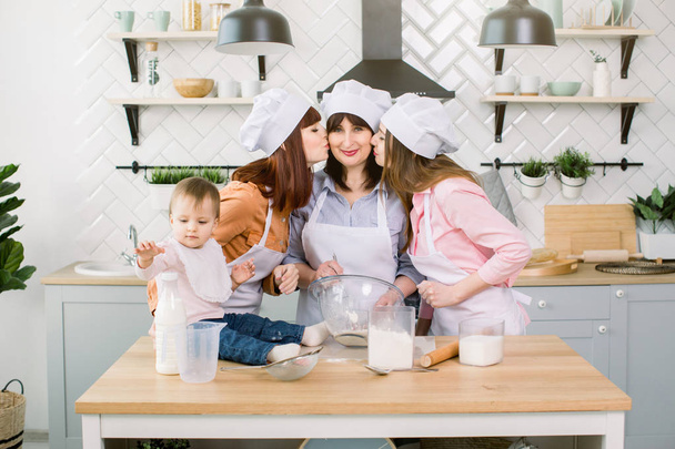 Two young sisters, grandmother and little baby daughter having good time in the kitchen. Daughters are kissing their mother - Foto, Imagen