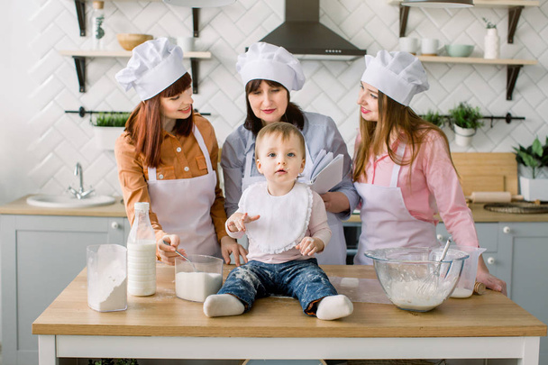 Little baby girl is sitting on the wooden table at kitchen while her mother, aunt and grandmother read the book with recipes on the background. Happy women in white aprons baking together - Φωτογραφία, εικόνα