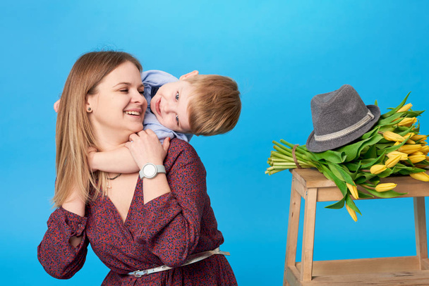 Portrait of a young mother and small toddler son having fun. Nearly lies a bouquet of spring yellow flowers, tulips. Family, adorable kid, love and happiness concept - Fotoğraf, Görsel