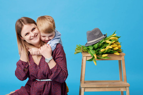 Portrait of a young mother and small toddler son having fun. Nearly lies a bouquet of spring yellow flowers, tulips. Family, adorable kid, love and happiness concept - Photo, Image