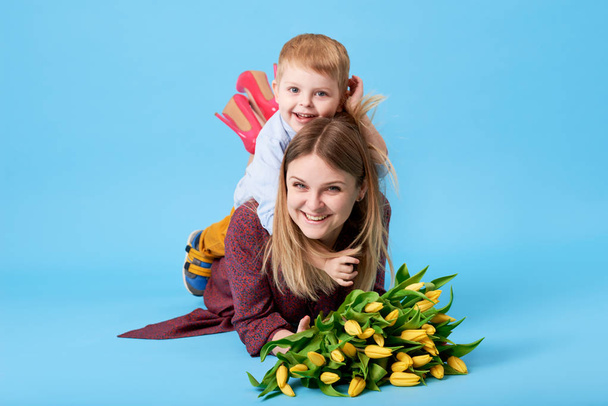 Portrait of a young mother and small toddler son having fun. Nearly lies a bouquet of spring yellow flowers, tulips. Family, adorable kid, love and happiness concept - Foto, Bild