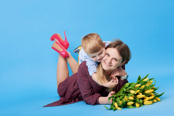 Portrait of a young mother and small toddler son having fun. Nearly lies a bouquet of spring yellow flowers, tulips. Family, adorable kid, love and happiness concept - Photo, Image