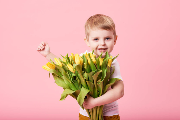 Adorable smiling child with spring flower bouquet looking at camera isolated on pink. Little toddler boy holding yellow tulips as gift for mom. Copy space for text  - Photo, Image