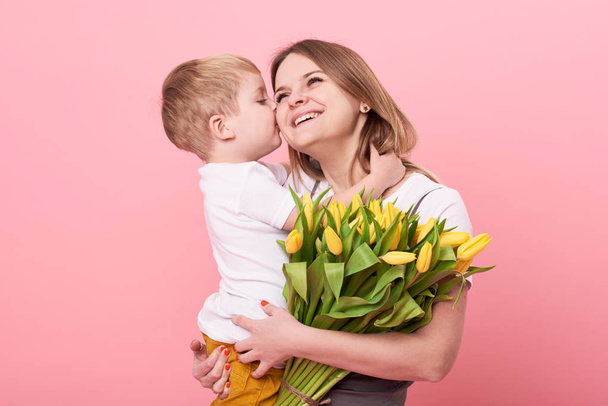 Young mother hugs his little son sitting on the floor against a pink background. Mom holds a bouquet of spring yellow flowers. Care and relationships and family concept. Mothers day - Foto, immagini