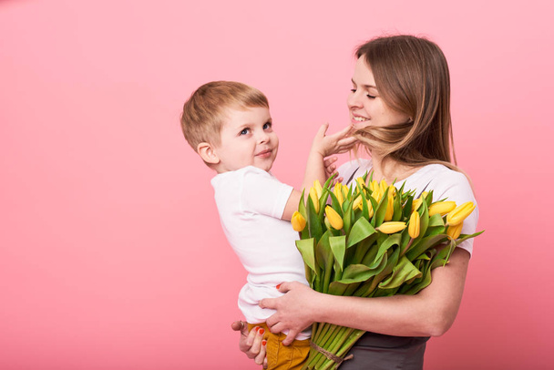 Mladá maminka objímá jeho malý syn sedí na podlaze růžové pozadí. Máma má kytice jarní žlutá. Koncept péče a vztahy a rodinu. Den matek - Fotografie, Obrázek