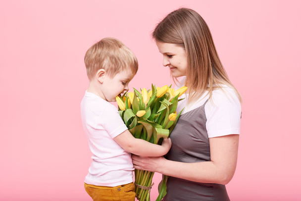 Jonge moeder knuffels zijn zoontje zittend op de vloer tegen een roze achtergrond. Moeder heeft een boeket van gele Lentebloemen. Zorg, relaties en familie concept. Moederdag - Foto, afbeelding