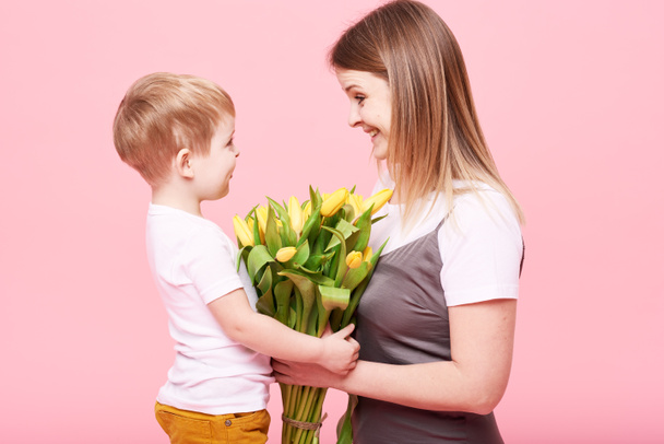 Madre joven abraza a su pequeño hijo sentado en el suelo sobre un fondo rosa. Mamá tiene un ramo de flores amarillas de primavera. Cuidado y relaciones y concepto familiar. Día de las madres
 - Foto, Imagen