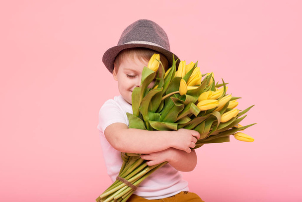 Adorable smiling child with spring flower bouquet looking at camera isolated on pink. Little toddler boy in hat holding yellow tulips as gift for mom. Copy space for text  - Photo, Image
