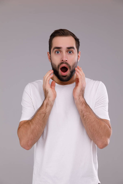 a man with a beard in a white T-shirt on a grey background surprised - Φωτογραφία, εικόνα