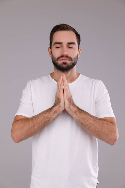 een man met een baard in een wit T-shirt op een grijze achtergrond bidden - Foto, afbeelding