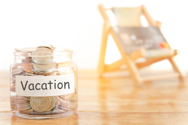 Vacation budget concept. Holidays money savings concept. Collecting money in the money jar for Vacation. Money jar with coins and beach seat on wooden background. - Photo, Image