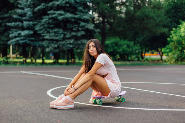 Portrait of a smiling charming brunette female sitting on her skateboard on a basketball court. - Foto, Imagem