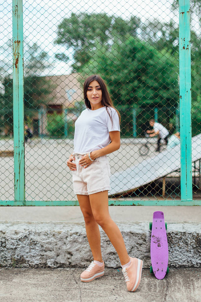Summer lifestyle image of trendy pretty young girl standing next to the scateboard coart with her plastic scateboard. - Φωτογραφία, εικόνα