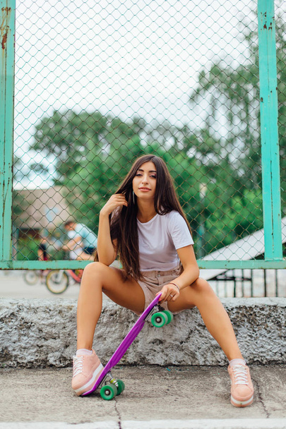 Summer lifestyle image of trendy pretty young girl sitting next to the skateboard coart with her plastic skateboard. - Fotó, kép