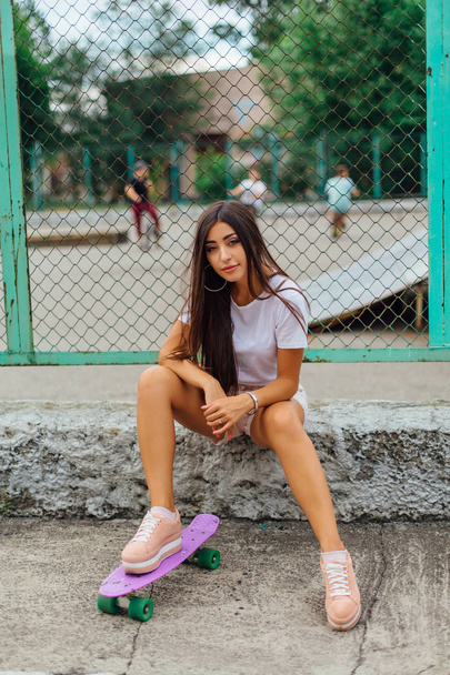 Summer lifestyle image of trendy pretty young girl sitting next to the skateboard coart with her plastic skateboard. - Φωτογραφία, εικόνα