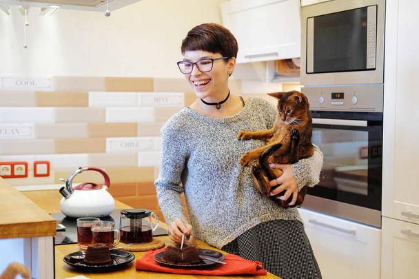 Hymyilevä nainen leikkii punapäisen kissan kanssa ja pelleilee keittiössä hänen kanssaan. Muotokuva naisen kotiäiti ruoanlaitto keittiössä
. - Valokuva, kuva