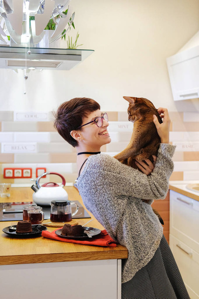 Mosolygó nő játszik a gyömbér cat, átölelve hoznak, és bolondozás körül a konyhában vele.  - Fotó, kép
