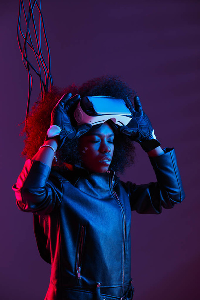Curly dark haired girl dressed in a black leather jacket and gloves is wearing  the virtual reality glasses on her head in the dark studio with neon light - Photo, Image