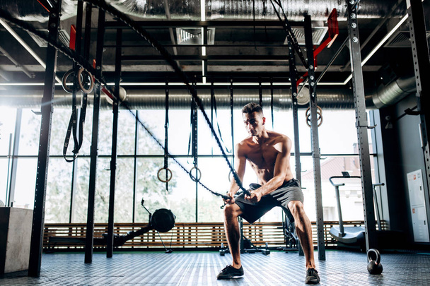Athletic dark-haired man with a naked torso dressed in the  black shorts doing sport exercises with sports ropes in the gym - Фото, зображення
