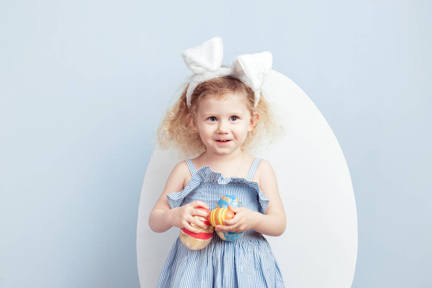 Charming curly girl in the light-blue dress with bunny ears on her head holds the dyed eggs in her hands on the background of a big white egg on a blue wall. Easter bunny - Foto, imagen