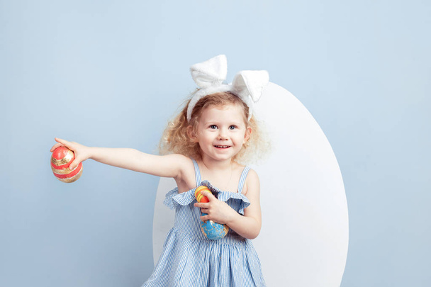 Charming curly girl in the light-blue dress with bunny ears on her head holds the dyed eggs in her hands on the background of a big white egg on a blue wall. Easter bunny - Фото, изображение